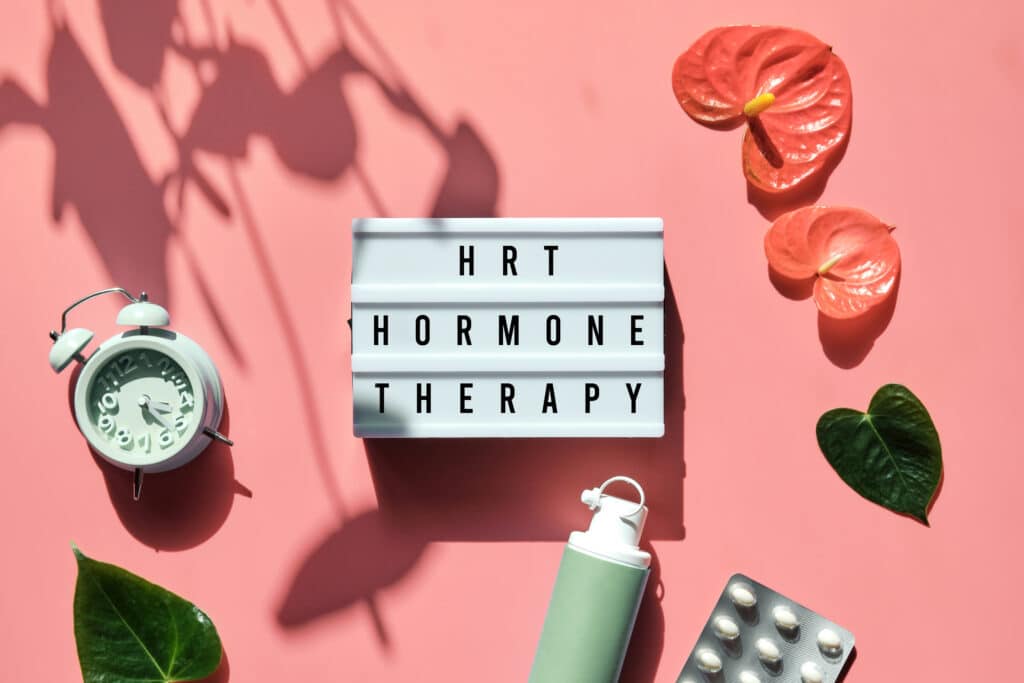Text HRT Replacement Therapy on light box. Menopause, hormone therapy concept. Oestrogen replacement therapy awareness. Pink background with alarm clock, exotic leaves, pills, estrogen gel.